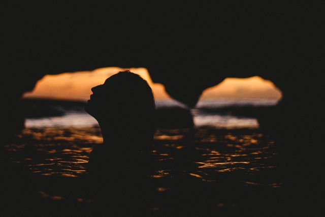Silhouette of woman swimming in ocean cave at sunset, casting an ethereal and serene ambiance. Perfect for themes conveying tranquility, nature, exploration, and the beauty of the outdoors. Great for use in travel advertisements, nature documentaries, wellness-themed projects, or inspirational and motivational content.