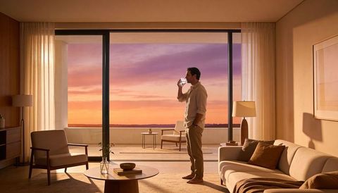 Man standing drinking water watching vibrant sunset over calm sea from modern suite balcony