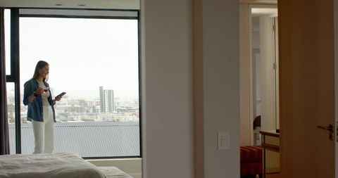 Female Traveler by Window with Smartphone and Coffee Mug in Modern Bedroom