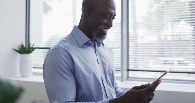 Smiling african american businessman using digital tablet and standing by window in office. business professional and working in busy modern office.