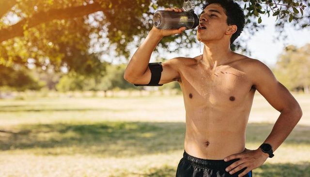 Shirtless young man hydrating after run in sunlit park with smartwatch and armband