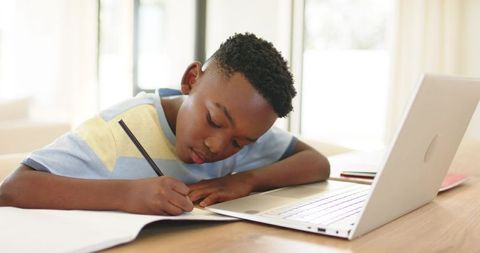 African American boy studying and writing homework at laptop on home desk in daylight