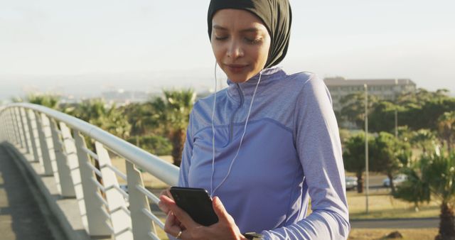 Smiling biracial woman wearing hijab, earphones and sports clothes using smartphone on city bridge. City living, communication, fitness and healthy modern urban lifestyle.