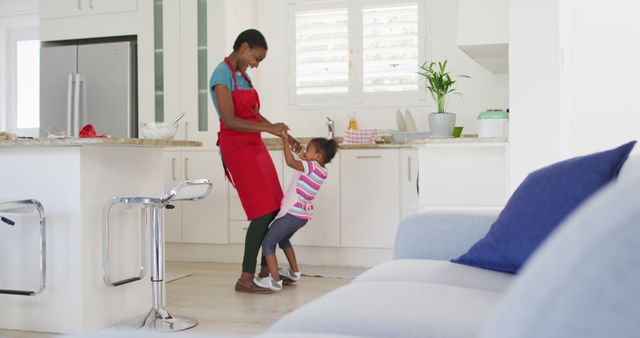 Mother and daughter sharing a joyful moment dancing in a modern kitchen with white cabinets and a bright atmosphere. The mother is wearing an apron, suggesting they might be cooking or baking together. This image highlights family bonding, parental affection, and the happiness of spending quality time indoors. Suitable for use in family lifestyle blogs, parenting articles, advertisements for household products, and home decor websites.