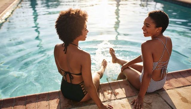 Two black women relaxing and splashing at sunlit poolside brick coping wearing swimsuits