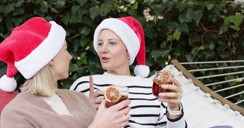 Friends wearing santa hats clinking glasses on hammock in backyard garden holiday cheers