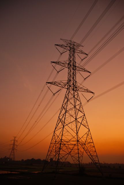 Silhouetted electricity transmission towers under a beautiful orange sky at dusk. Ideal for illustrating concepts related to energy infrastructure, power supply, technology, and sustainable energy. Can be used in articles about electricity, power grids, and advancements in energy transmission.