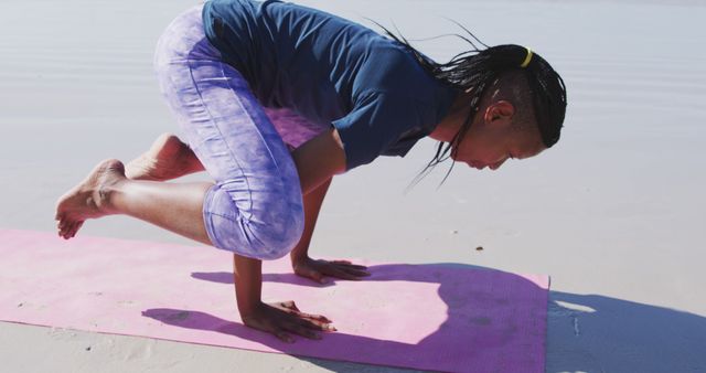 African American woman practicing yoga on a beach while performing a balance pose on pink yoga mat. Ideal for projects on fitness, wellness, relaxation, mental health, or outdoor activities.