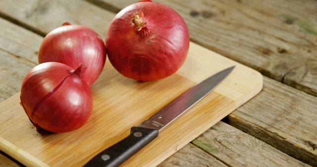 Red onions and knife placed on a wooden cutting board in a rustic kitchen. Perfect for use in cooking blogs, food preparation tutorials, recipe books, and advertisements related to fresh ingredients and kitchen tools.