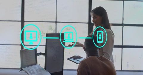 Asian woman pointing and gesturing to coworker at office desk with tablet and monitors