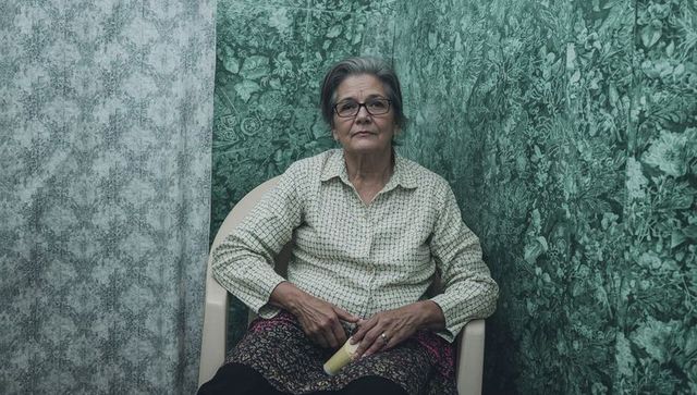 Elderly woman sitting in corner holding rolled paper surrounded by patterned wallpaper