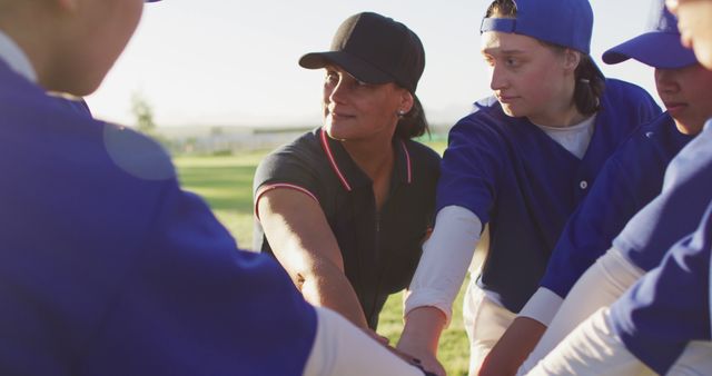 Female coach in black cap and sleeveless vest, encouraging a youth baseball team of boys wearing blue jerseys, standing together in a team huddle on a sunny outdoor playing field. Ideal for use in articles on team building, youth sports, coaching techniques, and female empowerment in sports.