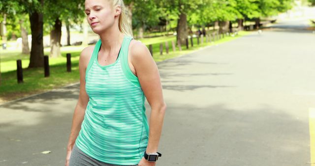 Athletic woman is taking a break during her workout in a park, showing signs of fatigue. She is dressed in a green tank top and wears a smartwatch. The scene is set in a tree-lined park under bright daylight. Ideal for themes related to fitness, healthy lifestyle, outdoor activities, and the realistic nature of physical exercise.