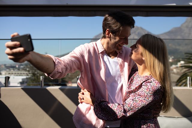 Front view of a happy Caucasian couple enjoying time off, on the roof terrace at a hotel on a sunny day, standing together embracing and smiling at each other, the man taking a selfie of them with a smartphone