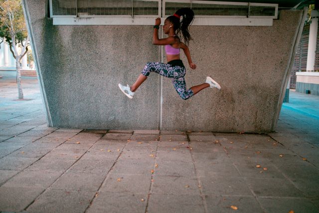 African American woman in athletic wear jumping under a bridge in an urban setting. Ideal for promoting fitness, active lifestyle, outdoor workouts, and health-related content. Suitable for use in advertisements, fitness blogs, and social media campaigns focused on exercise and motivation.