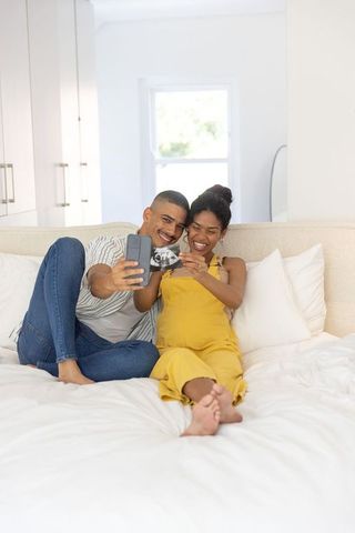Expectant couple smiling and sharing ultrasound while taking selfie on sunlit bed