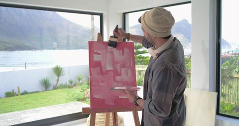 Indian artist painting abstract pink canvas in sunlit seaside studio with headphones and beanie