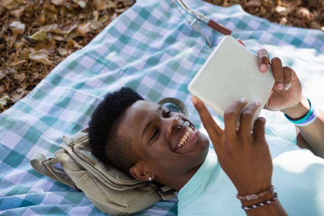 Man lying on a picnic blanket in a park, using a digital tablet and smiling. Ideal for promoting outdoor activities, technology usage in nature, leisure time, and lifestyle content. Suitable for articles, blogs, and advertisements focusing on relaxation, technology, and outdoor enjoyment.