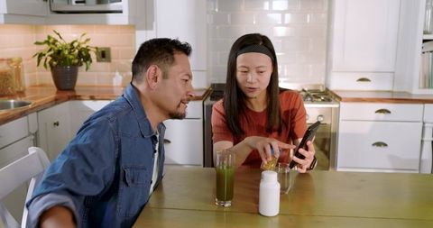 Asian couple researching supplement while pouring vitamins into glass in cozy kitchen