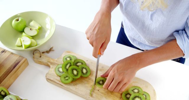 A person is slicing kiwi fruit on a wooden cutting board, with copy space. Fresh green apples are also visible in a bowl, indicating a setting of healthy food preparation.