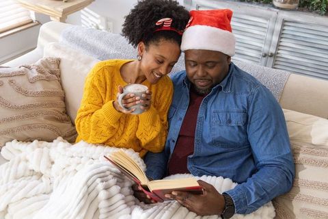 Cozy african american couple reading together on sofa wearing santa hat and holding mug