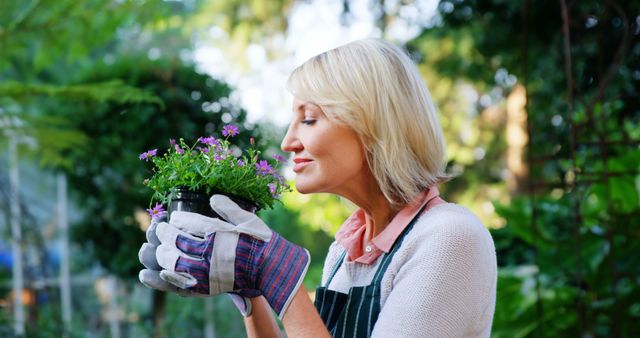 Mature woman enjoying gardening activities, smelling fresh flowers while wearing protective gardening gloves. Ideal for use in content related to outdoor hobbies, gardening tips, healthy living, relaxation in nature, and senior activities.