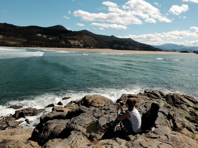 Man sitting on rocky coastline overlooking expansive ocean and distant mountains under clear sky with scattered clouds. Perfect for travel blogs, outdoor adventure promotions, nature retreats, or articles on solitude and relaxation.