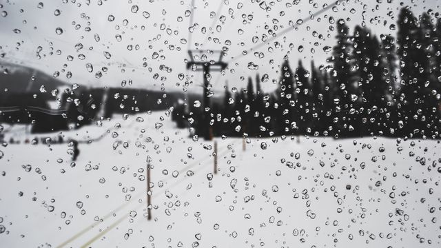 Snow-covered landscape viewed through a window with many droplets in focus. Snowy forest and ski lift blurred in the background. Perfect for winter, travel, and mood-themed uses.