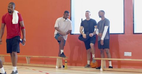 Diverse male basketball teammates greeting, stretching and preparing on indoor gym bench