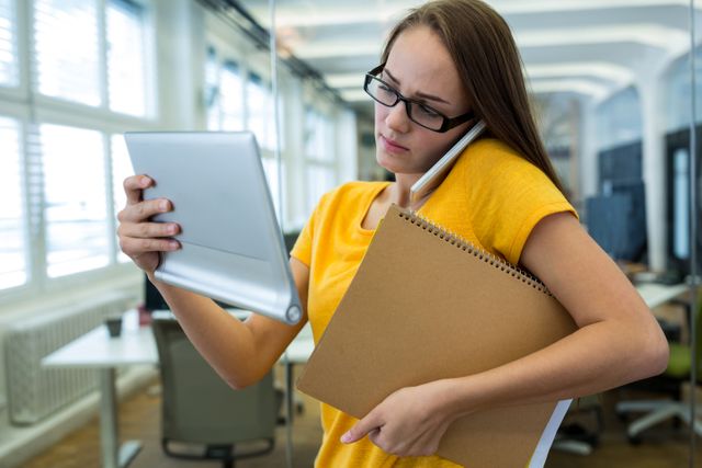 Female executive multitasking in a modern office, using a digital tablet and talking on a mobile phone while holding a notebook. Ideal for illustrating concepts of multitasking, productivity, modern business environments, and professional women in the workplace.