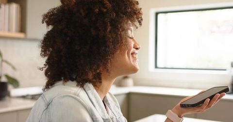 African American woman smiling while speaking into smartphone at modern kitchen counter