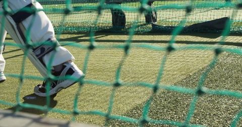 Batsman practicing footwork in net wearing batting pads on sunlit artificial turf
