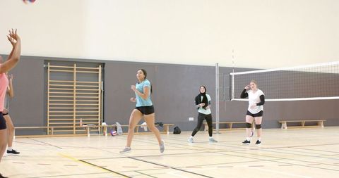 Diverse female teammates practicing indoor volleyball drill in gym with net and kneepads