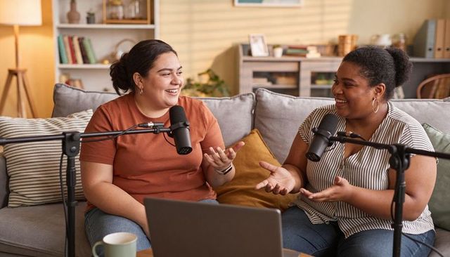 Two co-hosts recording candid podcast conversation in cozy home studio with laptop mics