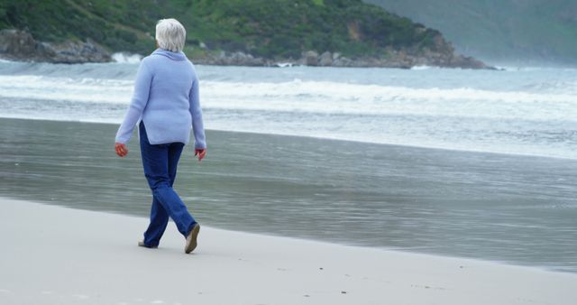 Senior woman walking alone on beach wearing winter clothing and blue sweater. Perfect for themes around solitude, reflection, retirement, active lifestyle for the elderly, and peaceful seaside moments. Great for use in articles or websites related to mental health, senior living, and nature walks.