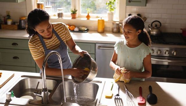 Mother daughter washing dishes together in sunlit kitchen teaching home cooking chores