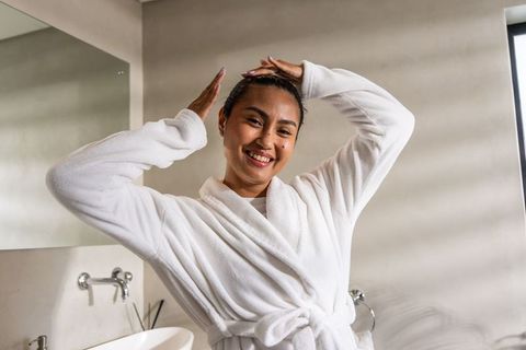 Smiling asian woman adjusting hair in plush white robe in minimalist bathroom morning routine
