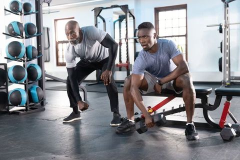 African american senior trainer coaching young man lifting hex dumbbell during strength workout