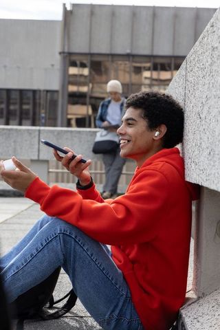 Teenager in red hoodie sitting on rooftop terrace using smartphone and wireless earbuds