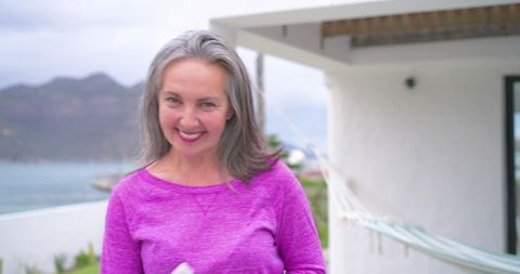 Smiling mature woman on coastal terrace wearing magenta top holding cloth near hammock