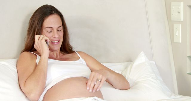 Smiling caucasian pregnant woman holding her belly using smartphone at home, copy space. Pregnancy, motherhood, domestic life and wellbeing concept, unaltered.