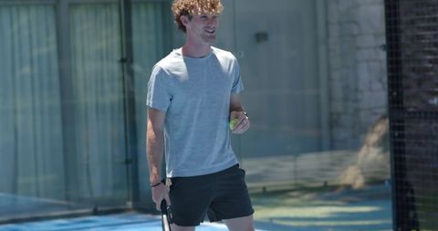 Man walking on glass-walled padel court holding racket and tennis ball, casual athletic