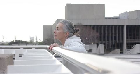 Contemplative mature woman leaning on rooftop railing in white hoodie overlooking skyline