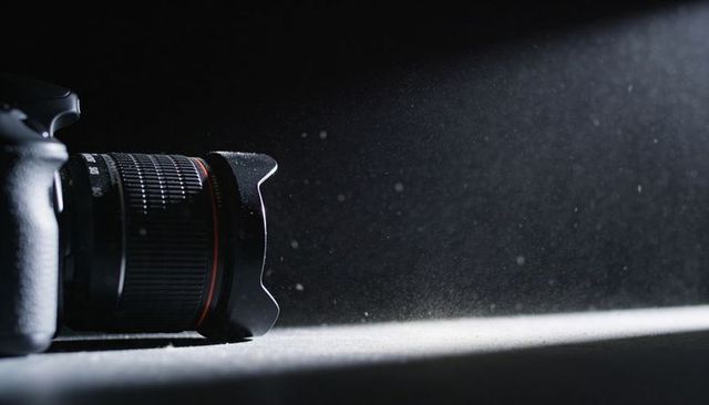 Pro camera lens with petal hood capturing dust beam in dramatic low-key studio composition