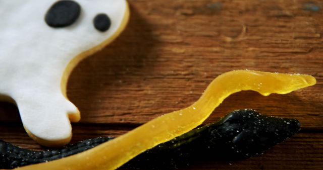 Close-up of a Halloween-themed cookie shaped like a ghost, paired with a gummy snake on a rustic wooden table. Perfect for use in holiday recipes, seasonal decorations, or children's party promotions.