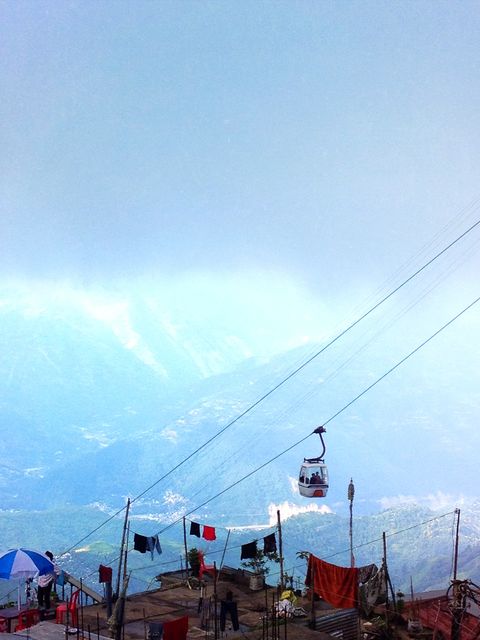 A scenic mountain cable car dangles high over a misty, mountainous landscape, with various human activities visible at the top of nearby buildings. Ideal for travel and adventure brochures, cultural exploration articles, or websites promoting remote destinations or hilly terrains. Icons signify transportation modes and unspoiled nature.