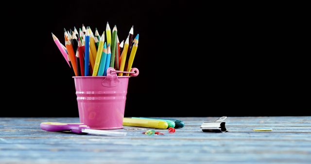 A pink bucket filled with colorful pencils is placed on a wooden surface, with copy space. Art supplies like this are often associated with creativity, education, and artistic endeavors.