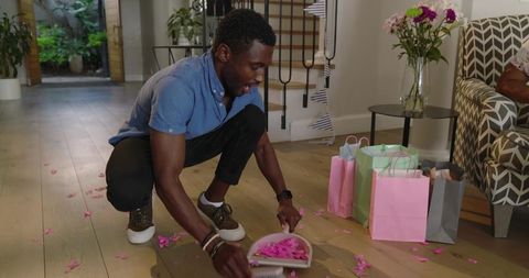 African American man sweeping pink petals near pastel gift bags in cozy living room