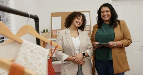 Women designers smiling while holding tablet in boutique fashion studio with garments