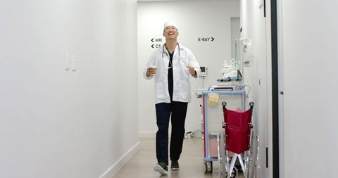 Asian doctor walking hospital corridor holding coffee cup and smiling with stethoscope
